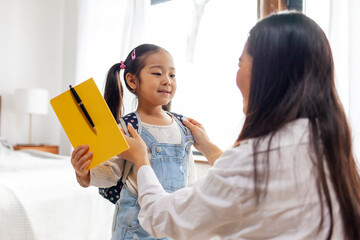 Wall Mural - mother collects Asian daughter to school, woman puts on backpack with book to little girl, back to school