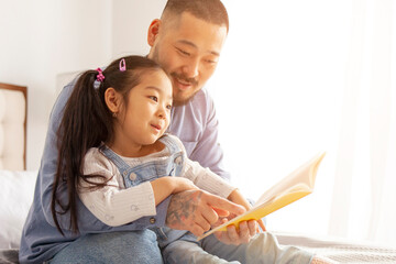 Wall Mural - little asian girl learn to read at home with dad, korean man with daughter sit on bed and learn lessons and homework