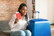 © Prostock-studio - Cheery african american woman sitting next to suitcase, holding passport and plane tickets, getting ready for trip