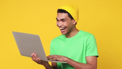 Wall Mural - Smart IT user young man of African American ethnicity wears green t-shirt hat hold in hand use work typing on laptop pc computer isolated on plain yellow background studio. People lifestyle concept