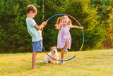 Fototapeta Zwierzęta - Children playing with pet dog jumping through hula hoop