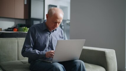 Elderly Man Using Laptop Sitting On Couch In Living Room At Home, Typing Text And Sending Message