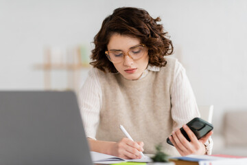 Wall Mural - pretty student in glasses holding smartphone while taking notes near blurred laptop on desk.