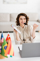 Wall Mural - happy language teacher in headset holding french dictionary while talking during online lesson near 
