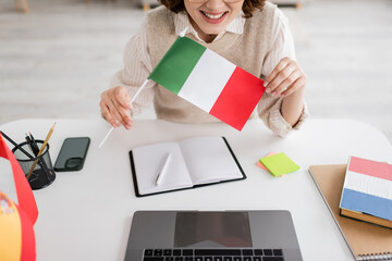 Wall Mural - cropped view of smiling language teacher holding italian flag near devices and notebooks on table at