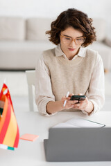 Wall Mural - young language teacher in glasses using smartphone near laptop and flags on blurred foreground.