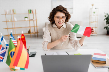 Wall Mural - young teacher holding flag of italy near laptop and textbook of french language while working at hom