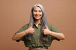 © Vadim Pastuh - Attractive middle-aged 50s woman with long grey hair showing thumbs up and looking at camera with toothy smile, lady agreeing with you, approving isolated on brown, good review