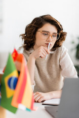 Wall Mural - pensive language teacher in glasses looking at laptop near international flags on blurred foreground