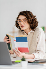 Wall Mural - curly student in glasses reading french dictionary book near laptop on desk.