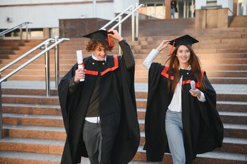 Wall Mural - Happy friends on graduation day. Portrait of two cheerful joyful students standing near university building.