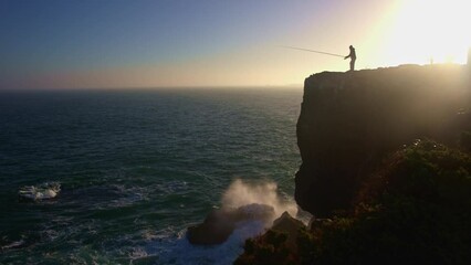 Wall Mural - A man stands on top of a cliff and fishes in the sea at sunset near by other men