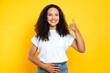 © Kateryna - Happy brazilian or hispanic woman in a white t-shirt, holds heone hand on her stomach and with another one shows thumb up gesture, depicting her good stomach health, smile, isolated yellow background