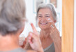 Portrait of happy senior beautiful woman applies anti aging cream on wrinkled face - elderly smiling lady looking in the mirror