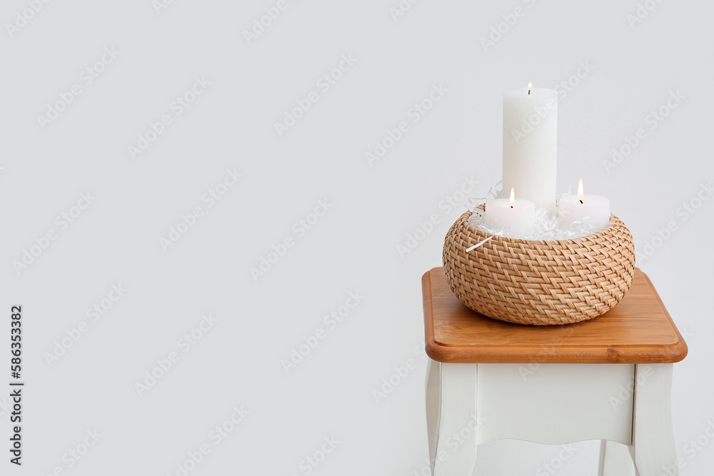 Basket with burning candles on table near light wall