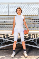 Wall Mural - Young teenage football player standing in front of sunny bleachers in football uniform holding football