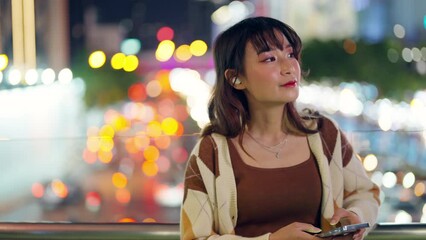 Wall Mural - Young Asian woman listening to the music on earphones and mobile phone application during travel in the city at night. Attractive girl have fun outdoor lifestyle using portable device with internet.