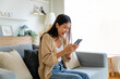 © Monster Ztudio - Happy young asian woman relax on comfortable couch at home texting messaging on smartphone, smiling girl use cellphone chatting, browse wireless internet on gadget, shopping online from home