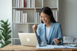 © crizzystudio - Asian businesswoman, investor showing a smile and Cheerful expression while looking at laptop receiving acceptance email Successful financial investments in office real estate business.