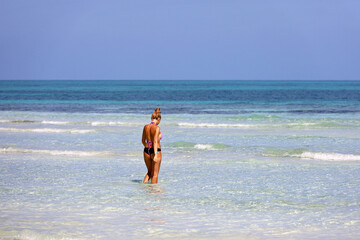 Tanned girl in swimsuit going to swim in blue sea water, back view. Beach vacation on Caribbean islands