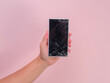 Close-up of hand holding mobile phone with broken touch screen on pink background. Female hand holding old phone with cracked and damaged screen.