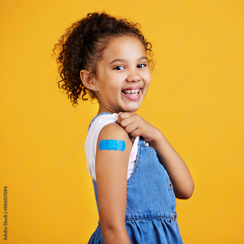 Happy, portrait and child with a plaster in a studio for a wound, sore or injury on her arm. Happiness, smile and healthy girl kid model posing with a bandaid after a vaccination by yellow background