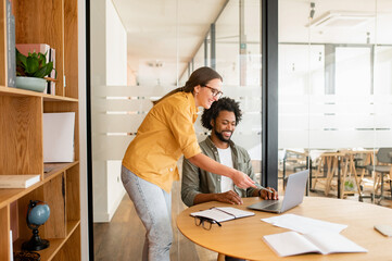 Pair of friendly colleagues work in office. Young guy and girl in glasses are working with a laptop in cosy office, a slight smile on their faces. Collaboration concept