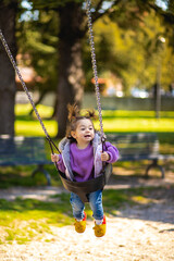 Wall Mural - Cute caucasian little toddler girl with two tails smiling widely and swinging on a baby swing in the playground with violet sweeter sleeveless jacket jeans