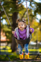Wall Mural - Sweet caucasian little toddler girl with two tails smiling widely and swinging on a baby swing in the playground with violet sweeter sleeveless jacket jeans