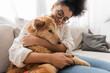 © LIGHTFIELD STUDIOS - Young african american woman in eyeglasses hugging dog on couch at home.