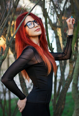 Wall Mural - Attractive young girl with red hair, wearing glasses, posing near a tree