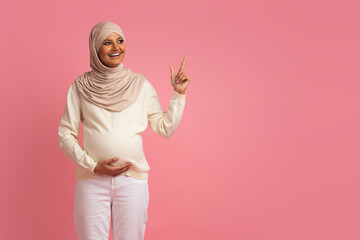 Wall Mural - Pregnant arab muslim woman touching belly and pointing aside at copy space