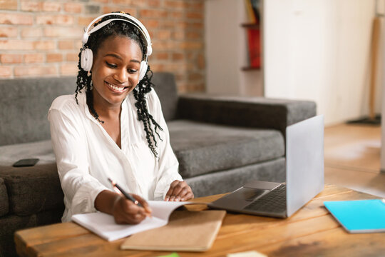 Wall Mural - Happy african american woman studying online from home, taking notes, sitting in front of laptop wearing headphones