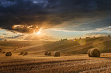 Fototapeta  - Stubble in a field with bales under a spectacular sky with clouds through which the sun's rays shine through