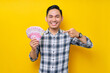 © Bangun Stock Photo - Portrait of smiling young Asian man wearing plaid shirt pointing finger at cash money in rupiah banknotes isolated on yellow background. people lifestyle concept
