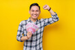 © Bangun Stock Photo - Portrait of excited young Asian man wearing plaid shirt holding cash money in rupiah banknotes and showing winner gesture isolated on yellow background. people lifestyle concept