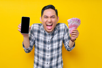 Wall Mural - Portrait of excited young Asian man wearing plaid shirt showing blank screen mobile phone and holding cash money in rupiah banknotes isolated on yellow background. people lifestyle concept