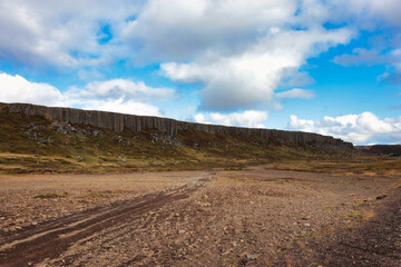 A vast, open landscape in Iceland with a rugged dirt path leading towards a rocky cliff under a bright blue sky with scattered clouds, showcasing the country's natural beauty and wilderness