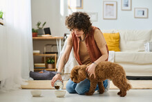 Young Woman Pouring Food Into Bowl And Feeding Her Dog In The Living Room At Home