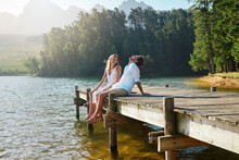 Young Couple, Lake And Jetty With Laughing, Happiness And Bonding With Love In Nature For Holiday. Man, Woman And Comic Joke On Bridge By Water To Relax With Conversation, Care And Vacation In Summer