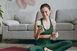 © ViDi Studio - Happy young smiling strong sporty athletic fitness trainer instructor woman wears green tracksuit hold use mobile cell phone drink water train do exercises at home gym indoor. Workout sport concept.