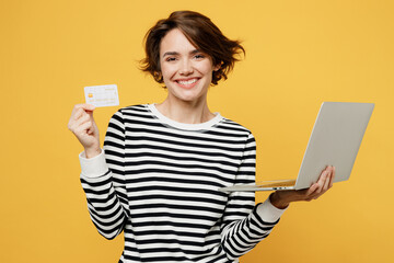 Wall Mural - Young fun IT woman wear casual striped shirt using mobile cell phone hold credit bank card doing online shopping order delivery booking tour isolated on plain yellow color background studio portrait.