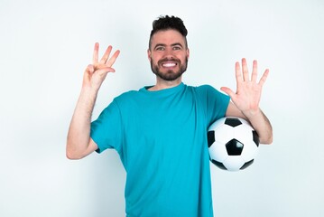 Sticker - Young man holding a ball over white background showing and pointing up with fingers number nine while smiling confident and happy.