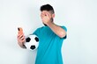 © Jihan - Young man holding a ball over white background looking at smart phone feeling sad holding hand on face.