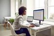 © Studio Romantic - Female accountant working on office computers. Young African American woman sitting at her desk, using desktop and laptop computers, looking at the screen, and working with business spreadsheets