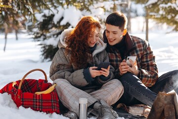  A young red-haired woman, along with her boyfriend, are watching something on their smartphones while sitting on a halt in a winter forest. A young couple had a picnic in the winter forest.