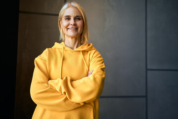 Portrait of a beautiful senior modern woman in a yellow hoodie, crossing her arms and smiling, looking at the camera.  Carefree and senior life.