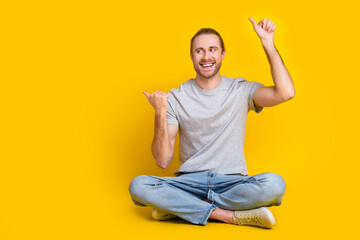 Sticker - Full size photo of positive man gray t-shirt pants sitting on floor look indicating empty space sale isolated on yellow color background