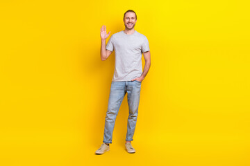 Sticker - Full size photo of good mood friendly man wear gray t-shirt jeans hand in pocket waving palm say hi isolated on yellow color background