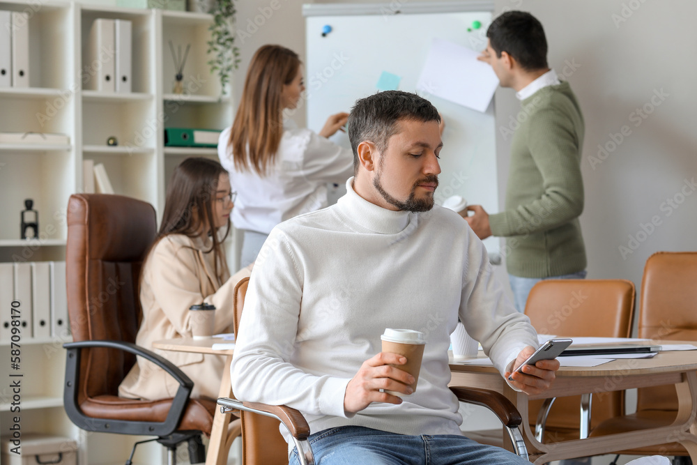 Handsome businessman with cup of coffee using mobile phone in office
